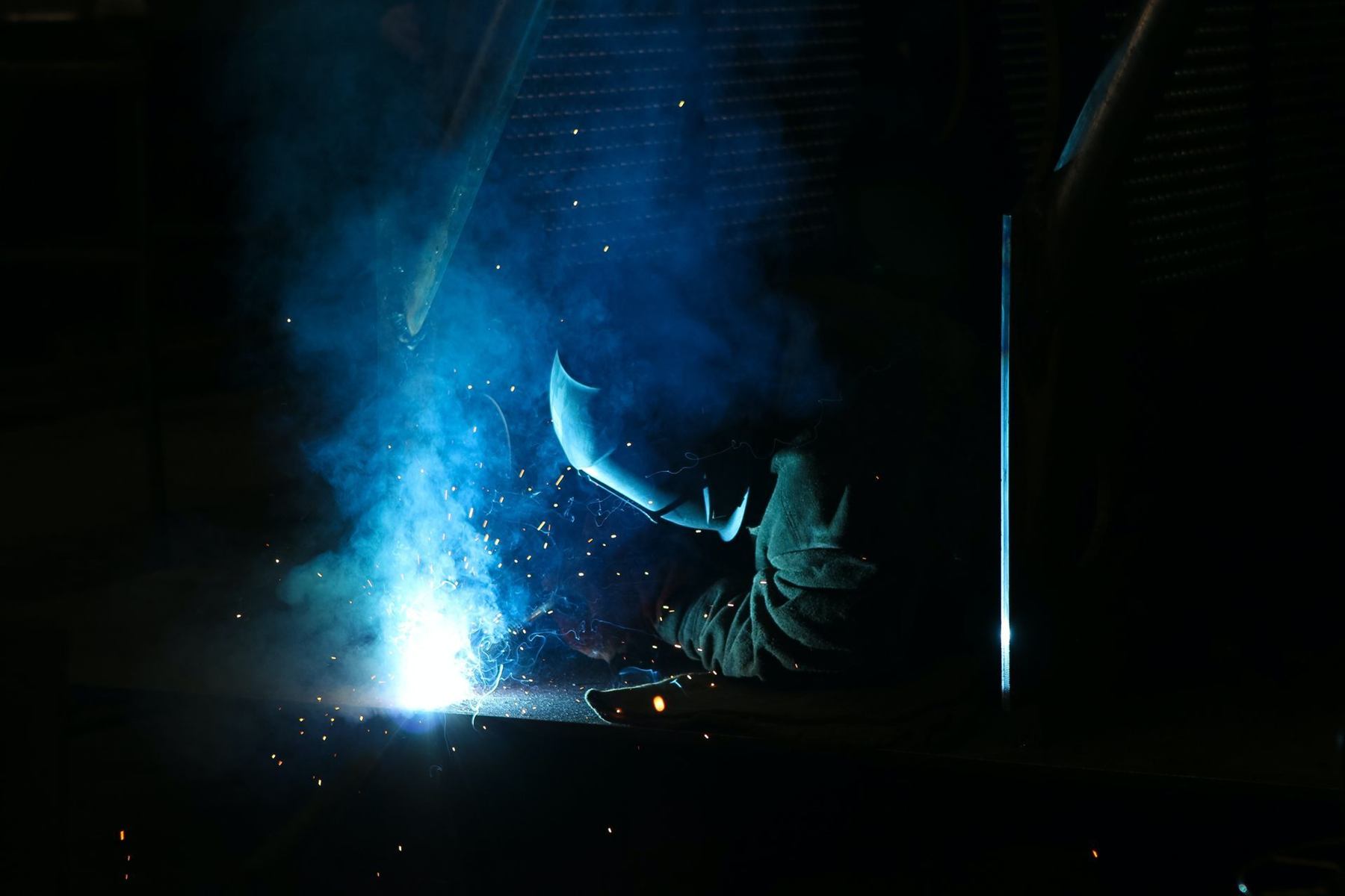 Welder at work in the dark — blue arc glow, smoke and orange sparks flying off the steel